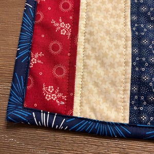 May include: A quilted placemat with a patriotic theme. It features red fabric with white floral patterns, a cream-colored star-patterned stripe, and blue fabric with white starburst designs. The edges are trimmed with a blue fabric.