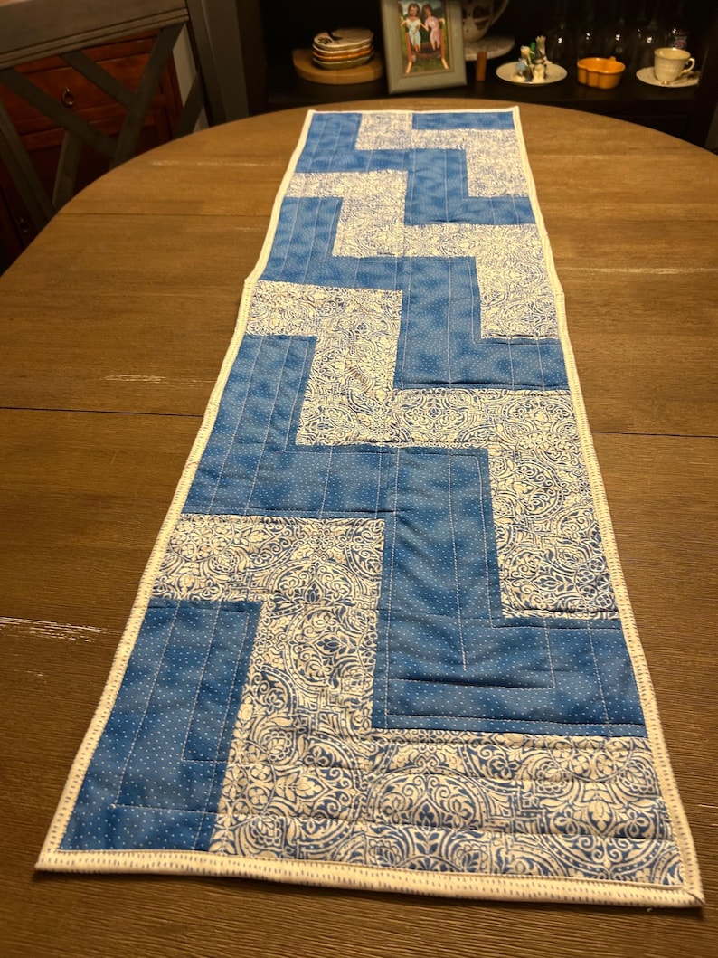 Pu&ograve; includere: Runner da tavolo trapuntato blu e bianco con un design geometrico. Il runner presenta blocchi alternati di tessuto blu tinta unita e tessuto bianco fantasia con accenti blu. I bordi sono rifiniti con un bordo bianco.