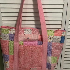Peut inclure: Sac fourre-tout matelassé avec un motif patchwork dans les tons de rose, vert, violet et blanc. Le sac a une grande poche avant et de longues anses roses. Le tissu présente des motifs floraux et d'insectes.