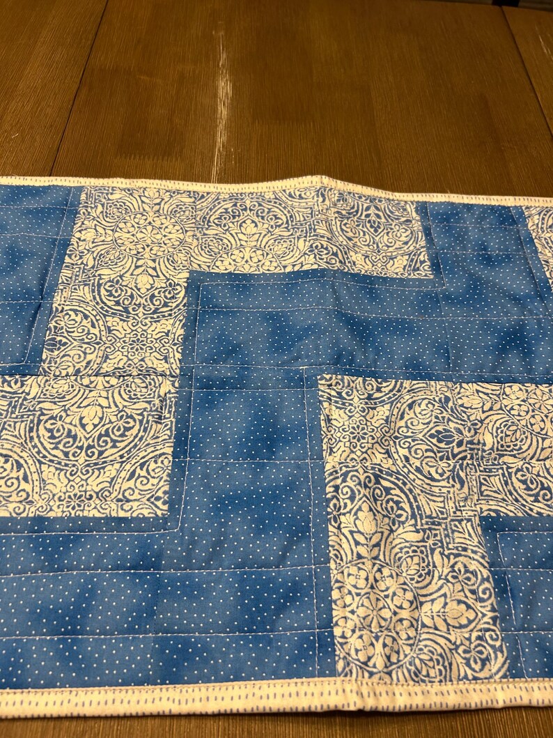 Pu&ograve; includere: Una tovaglia trapuntata blu e bianca. La tovaglia presenta un motivo di tessuto blu con pois bianchi e sezioni con un motivo floreale bianco. I bordi sono rifiniti con un bordo e cuciture bianche.