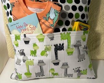 Pode incluir: Um travesseiro de tecido branco com um padrão de bolinhas verdes e pretas. O travesseiro tem um bolso na frente com uma estampa de girafas verdes e brancas. O bolso contém um livro intitulado "How Do Lions Say Wow?", uma camiseta laranja, um pijama e uma caixa de lápis de cor.