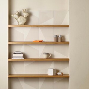 May include: A set of four wooden shelves with a white geometric tile background. The shelves are empty except for a white ceramic cactus sculpture, a small orange tray, two small gray bottles, a white ceramic mug, a white cube, and a stack of books.