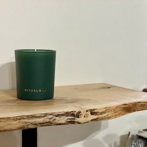 Könnte beinhalten: Eine grüne, gefrostete Glaskerze mit dem Text "RITUALS..." auf der Vorderseite, die auf einem Holzregal steht.