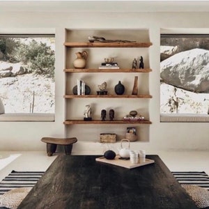 May include: A rustic wooden shelf unit with five shelves, featuring various decorative items, including a ceramic vase, a wooden sculpture, and a collection of books. The shelves are mounted against a white wall, with two large windows on either side.