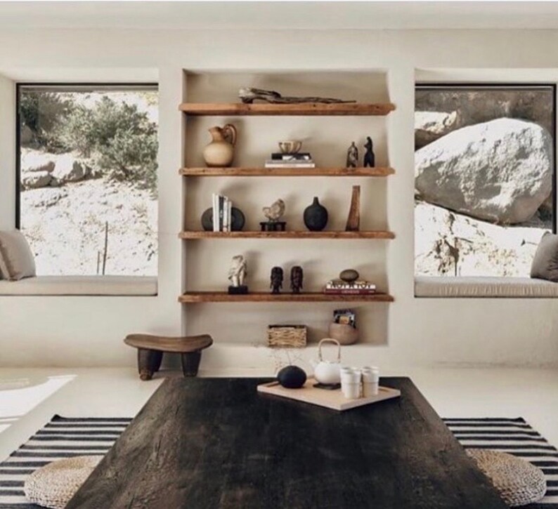 May include: A rustic wooden shelf unit with five shelves, featuring various decorative items, including a ceramic vase, a wooden sculpture, and a collection of books. The shelves are mounted against a white wall, with two large windows on either side.
