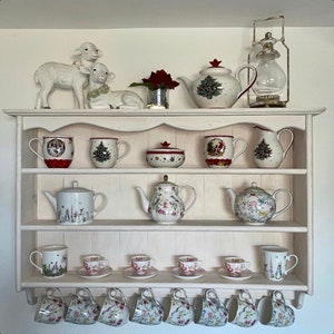 Peut inclure: Une étagère en bois blanc vieilli présentant diverses tasses à thé, théières, mugs et objets décoratifs. Les articles comprennent des mugs et théières sur le thème de Noël avec des motifs de sapins, ainsi que des tasses à thé et soucoupes à motifs floraux.