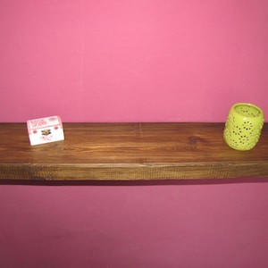 May include: A wooden shelf with a light brown finish. The shelf is mounted on a pink wall. A small white box with red and pink floral designs is on the left side of the shelf. A green candle holder with a floral design is on the right side of the shelf.