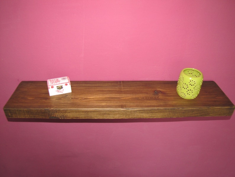 May include: A wooden shelf with a light brown finish. The shelf is mounted on a pink wall. A small white box with red and pink floral designs is on the left side of the shelf. A green candle holder with a floral design is on the right side of the shelf.
