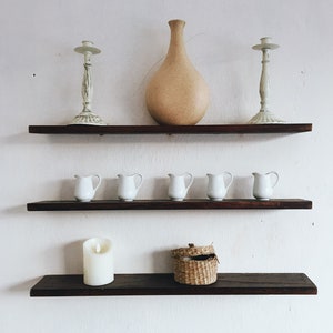 May include: Three wooden shelves with white ceramic candle holders, a tan ceramic vase, and white ceramic pitchers. The bottom shelf also has a white candle and a small woven basket.