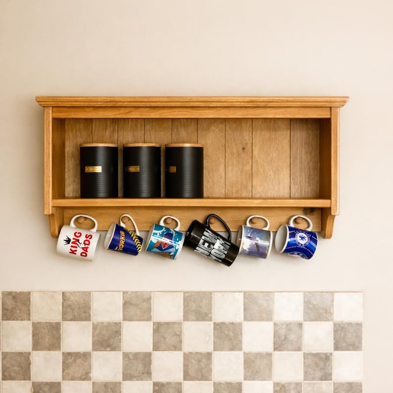 May include: A light brown wooden wall shelf with a top ledge and six hooks. Three black cylindrical canisters with wooden lids sit on the shelf. Six ceramic mugs hang from the hooks, featuring various designs including text reading KING DADS and NEW YORK.