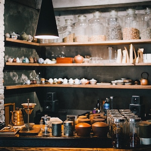 May include: A rustic shop interior with wooden shelves stocked with glass jars, teacups, and other tea-related items. The shelves are against a dark gray wall. There is a sign on the wall with Chinese characters that read "有為者, 必有其志".