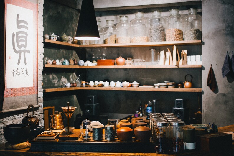 May include: A rustic shop interior with wooden shelves stocked with glass jars, teacups, and other tea-related items. The shelves are against a dark gray wall. There is a sign on the wall with Chinese characters that read "有為者, 必有其志".