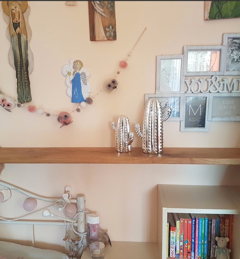 May include: A white shelf with two silver cactus figurines on it. The shelf is mounted on a light pink wall. There is a white picture frame with the text "You & Me" and a string of white and pink pom-poms hanging on the wall.