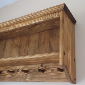 May include: A wall-mounted wooden shelf featuring a natural wood grain finish with a horizontal storage ledge and a lower section equipped with four protruding pegs for hanging items. The unit is constructed from stained pine wood planks.