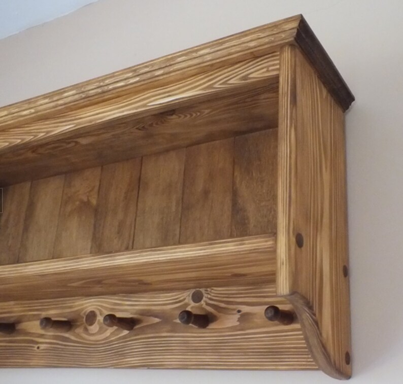 May include: A wall-mounted wooden shelf featuring a natural wood grain finish with a horizontal storage ledge and a lower section equipped with four protruding pegs for hanging items. The unit is constructed from stained pine wood planks.