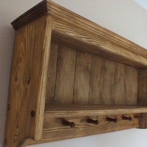 May include: A wall-mounted wooden shelf featuring a natural brown wood grain finish, a flat top ledge, and a lower horizontal bar equipped with five small, evenly spaced wooden pegs for hanging items, constructed with visible vertical wood paneling.