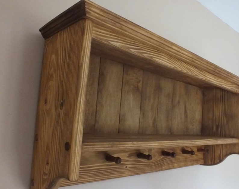 May include: A wall-mounted wooden shelf featuring a natural brown wood grain finish, a flat top ledge, and a lower horizontal bar equipped with five small, evenly spaced wooden pegs for hanging items, constructed with visible vertical wood paneling.