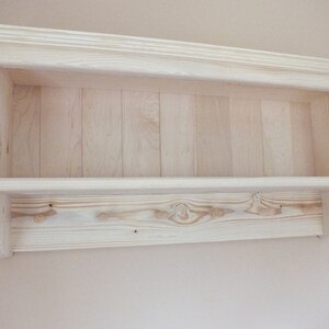 May include: A light-colored wooden wall shelf featuring a horizontal storage surface and a lower panel equipped with four integrated wooden pegs for hanging items. The unit has a crown molding top edge and a vertical plank backboard design.
