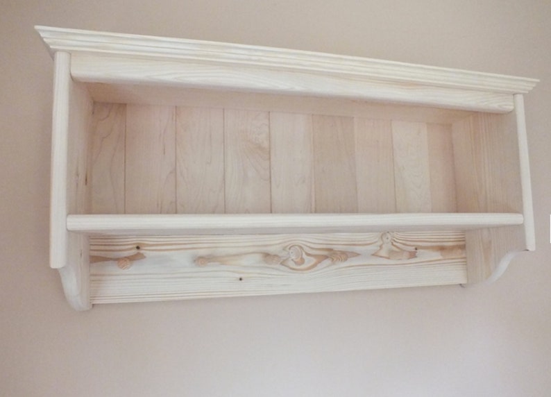 May include: A light-colored wooden wall shelf featuring a horizontal storage surface and a lower panel equipped with four integrated wooden pegs for hanging items. The unit has a crown molding top edge and a vertical plank backboard design.