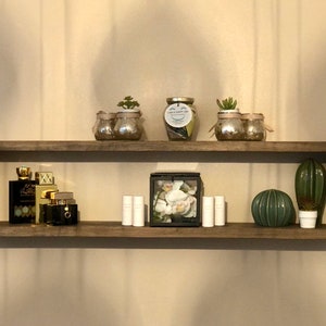 Puede incluir: Dos estantes de madera con varios artículos en ellos. El estante superior tiene tres frascos de vidrio con pequeñas plantas. El frasco del medio tiene una etiqueta blanca que dice "I am a happy jar". El estante inferior tiene tres botellas de perfume, tres pequeños tubos, una caja negra con flores blancas, un cactus verde y un cactus de vidrio verde.