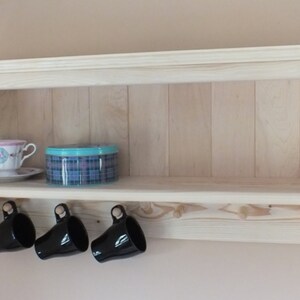 May include: A light-colored wooden wall shelf with a flat top surface and a lower rail featuring six wooden pegs. Three black ceramic mugs hang from the left pegs. A floral teacup on a saucer and a round blue plaid tin sit on the shelf.
