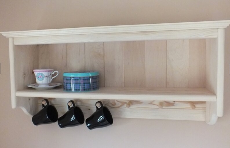 May include: A light-colored wooden wall shelf with a flat top surface and a lower rail featuring six wooden pegs. Three black ceramic mugs hang from the left pegs. A floral teacup on a saucer and a round blue plaid tin sit on the shelf.