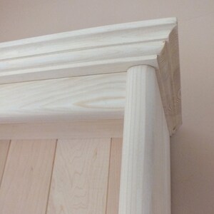 May include: A light-colored wooden shelf unit featuring a vertical cylindrical support pillar on the right side and a horizontal crown molding top piece with decorative grooves, showcasing the natural grain of the unfinished wood.