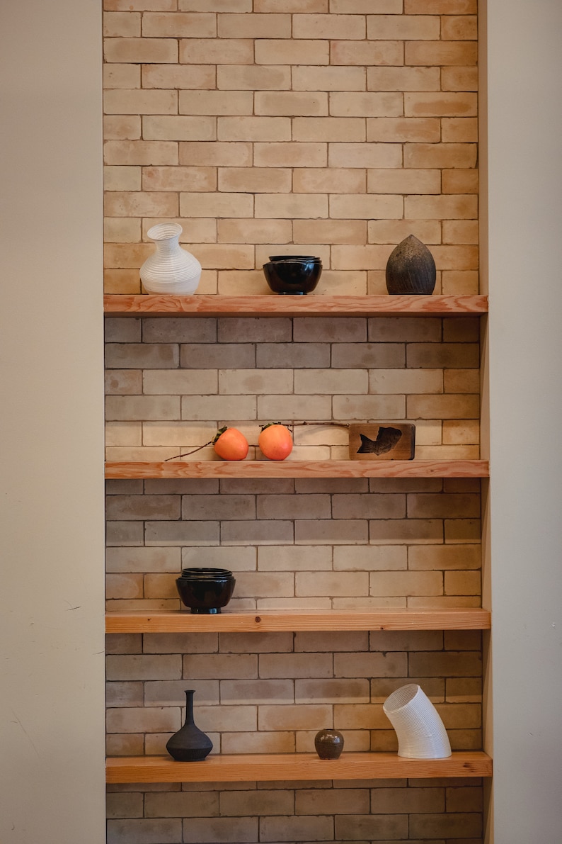 May include: A set of wooden shelves with various ceramic and wooden objects, including a white vase, a black bowl, a wooden sculpture, two orange persimmons, a black vase, and a white ceramic tube.