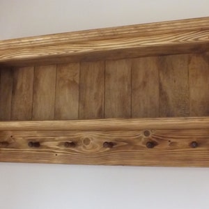 May include: A wall-mounted wooden shelf featuring a horizontal storage ledge and a lower panel equipped with six small, evenly spaced pegs for hanging items. The unit is constructed from light brown wood with a visible grain pattern.