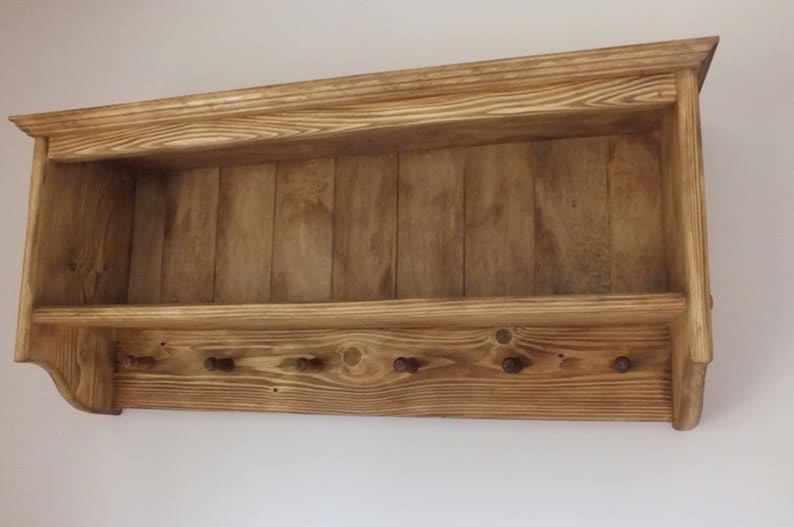 May include: A wall-mounted wooden shelf featuring a horizontal storage ledge and a lower panel equipped with six small, evenly spaced pegs for hanging items. The unit is constructed from light brown wood with a visible grain pattern.