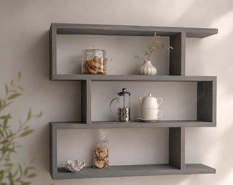 May include: A gray wooden wall shelf with a three-tiered, staggered, S-shaped design. The shelves hold a glass jar with cookies, a small white vase with a plant, a glass French press, a white teapot, a white teacup, and a small bowl.