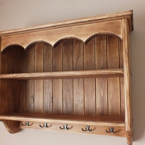 May include: A wooden wall shelf unit with two shelves and hooks. The light brown wood has a distressed finish. The top features an arched design. The unit is designed for wall mounting and has a rustic aesthetic.
