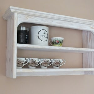 Peut inclure: Étagère murale en bois blanc à deux niveaux. L'étagère supérieure comporte un bocal en verre étiqueté "HOME", une tasse blanche avec le mot "KAWA" et un motif de papillon, ainsi qu'une tasse à thé blanche avec un motif rouge et vert. L'étagère inférieure comporte six tasses à thé blanches avec des motifs floraux.