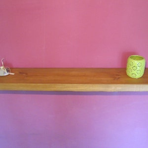 May include: A wooden shelf with a light brown finish, mounted on a pink wall. A small white object and a green candle holder are on the shelf.