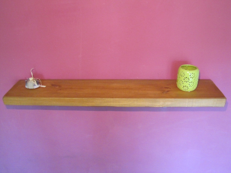 May include: A wooden shelf with a light brown finish, mounted on a pink wall. A small white object and a green candle holder are on the shelf.