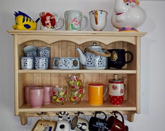May include: A light-colored wooden wall shelf with three tiers holding various mugs, teapots, and glassware. The top shelf features character-themed items, the middle shelf displays blue and white patterned ceramics, and the bottom shelf holds assorted colorful mugs and glasses, with additional mugs hanging from hooks.