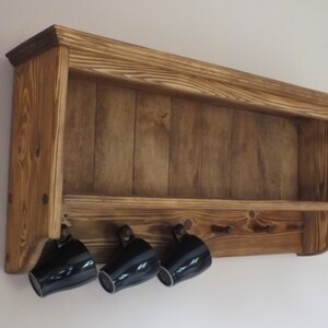 May include: A wall-mounted wooden shelf with a dark brown stained finish and visible wood grain. The unit features an open top storage area and a lower section with five small pegs, three of which hold black ceramic mugs hanging downward.