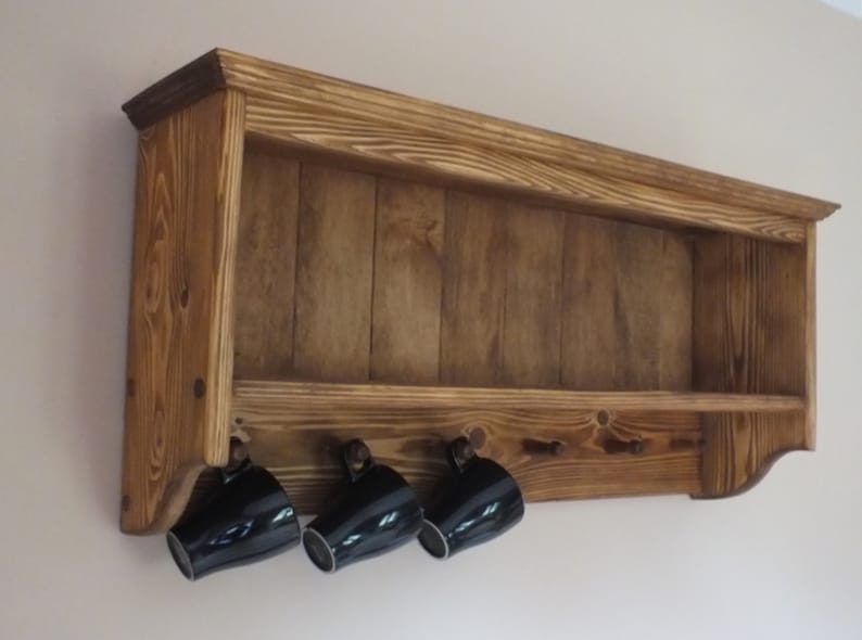 May include: A wall-mounted wooden shelf with a dark brown stained finish and visible wood grain. The unit features an open top storage area and a lower section with five small pegs, three of which hold black ceramic mugs hanging downward.