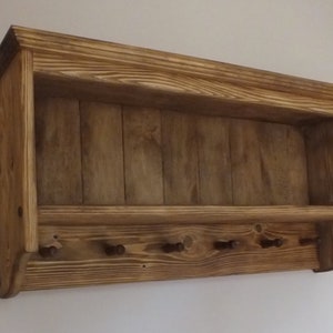 May include: A wall-mounted wooden shelf featuring a natural brown wood grain finish. The unit includes an upper storage ledge and a lower panel equipped with six small, evenly spaced wooden pegs for hanging items in a horizontal row.