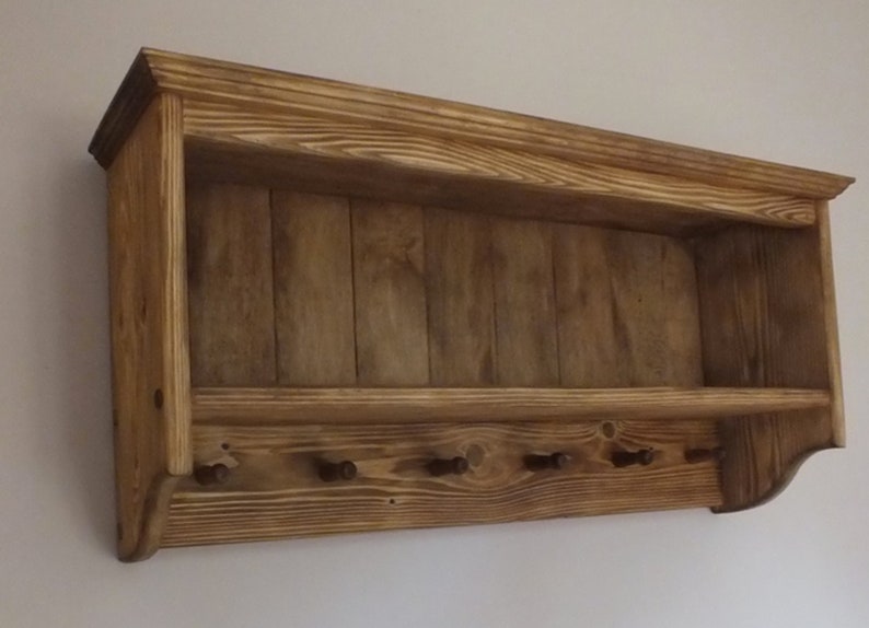 May include: A wall-mounted wooden shelf featuring a natural brown wood grain finish. The unit includes an upper storage ledge and a lower panel equipped with six small, evenly spaced wooden pegs for hanging items in a horizontal row.