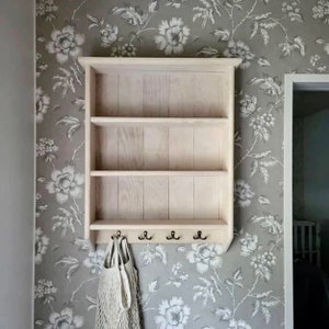 Op de afbeelding: Wit houten wandrek met drie planken en vier haken. Het rek is gemonteerd aan een muur met een bloemig behangpatroon.