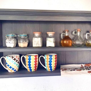 Könnte beinhalten: Ein dunkles graues Wandregal aus Holz mit zwei Ablagen. Die obere Ablage hat sechs Glasbehälter mit Deckeln, von denen drei orangefarbene Deckel haben. Die untere Ablage hat drei bunte Tassen mit geometrischen Mustern und ein weißes Tuch mit einem Blumenmuster.