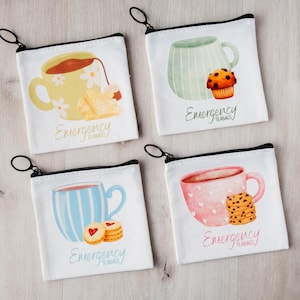 Peut inclure: Quatre pochettes en toile blanche avec des fermetures éclair et des tirettes noires, chacune avec une illustration à l'aquarelle d'une tasse et de collations. Les motifs incluent du thé, un muffin, des biscuits et le texte "Emergency Teas!"