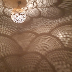 May include: A round, white, metal light fixture with a decorative pattern casts intricate shadows on a beige ceiling. The light fixture is hanging from the ceiling.