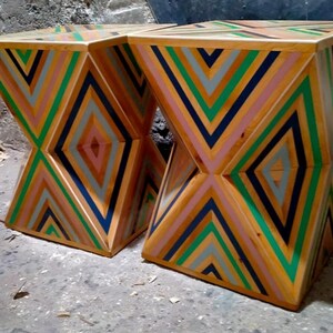 K&ouml;nnte beinhalten: Zwei Holzhocker mit einem geometrischen Muster aus rosa, gr&uuml;nen, blauen und grauen Streifen. Die Hocker sind aus Holz gefertigt und haben ein einzigartiges, modernes Design.
