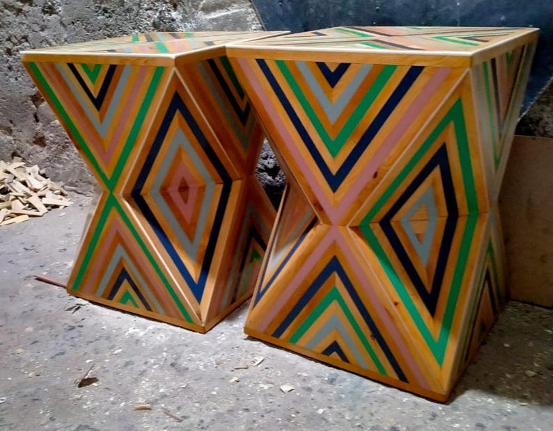 K&ouml;nnte beinhalten: Zwei Holzhocker mit einem geometrischen Muster aus rosa, gr&uuml;nen, blauen und grauen Streifen. Die Hocker sind aus Holz gefertigt und haben ein einzigartiges, modernes Design.