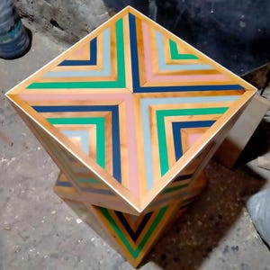 K&ouml;nnte beinhalten: Ein quadratischer Holzhocker mit einem geometrischen Muster auf der Oberseite. Das Muster besteht aus abwechselnden Streifen aus rosa, blau, gr&uuml;n und braunem Holz. Der Hocker ist auf einem anderen &auml;hnlichen Hocker gestapelt.