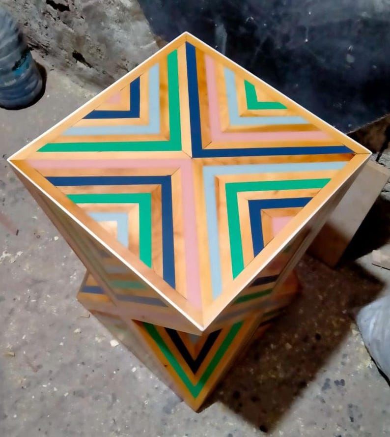 K&ouml;nnte beinhalten: Ein quadratischer Holzhocker mit einem geometrischen Muster auf der Oberseite. Das Muster besteht aus abwechselnden Streifen aus rosa, blau, gr&uuml;n und braunem Holz. Der Hocker ist auf einem anderen &auml;hnlichen Hocker gestapelt.