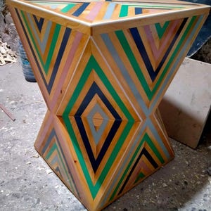 K&ouml;nnte beinhalten: Ein geometrischer Holzhocker mit einem einzigartigen Design mit abwechselnden Streifen in Gr&uuml;n, Schwarz, Rosa und Grau. Der Hocker hat eine quadratische Oberseite und eine rautenf&ouml;rmige Basis.