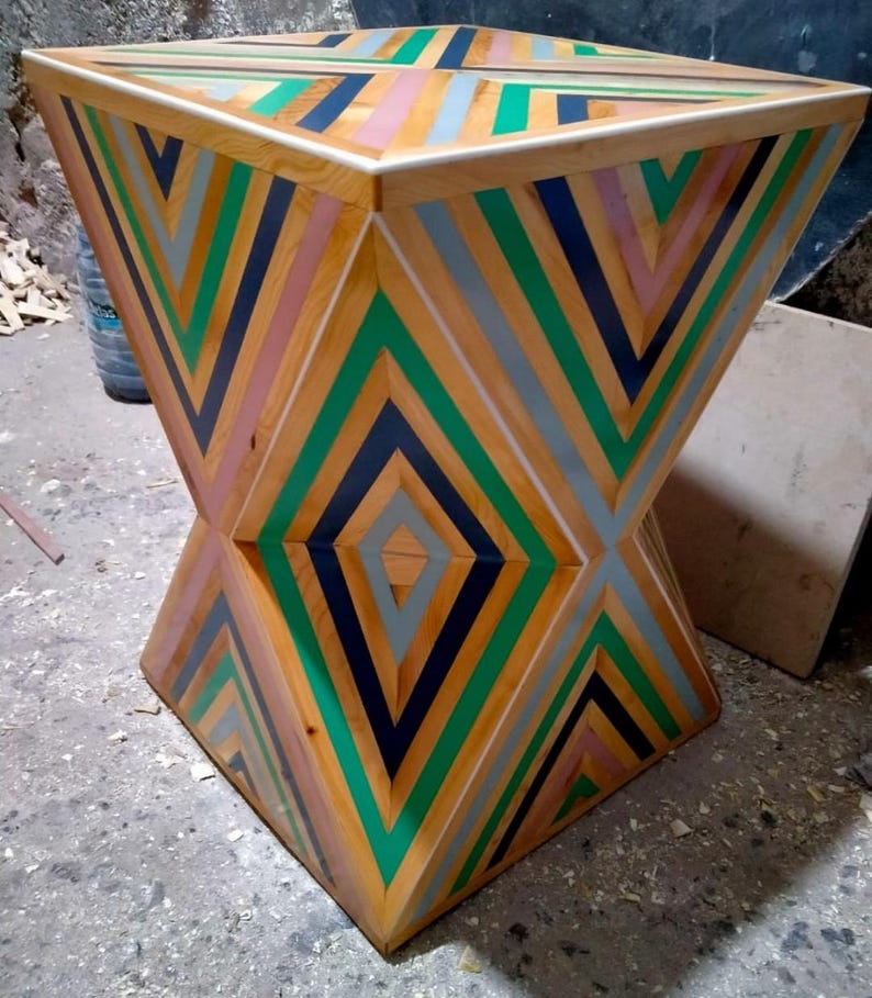 K&ouml;nnte beinhalten: Ein geometrischer Holzhocker mit einem einzigartigen Design mit abwechselnden Streifen in Gr&uuml;n, Schwarz, Rosa und Grau. Der Hocker hat eine quadratische Oberseite und eine rautenf&ouml;rmige Basis.